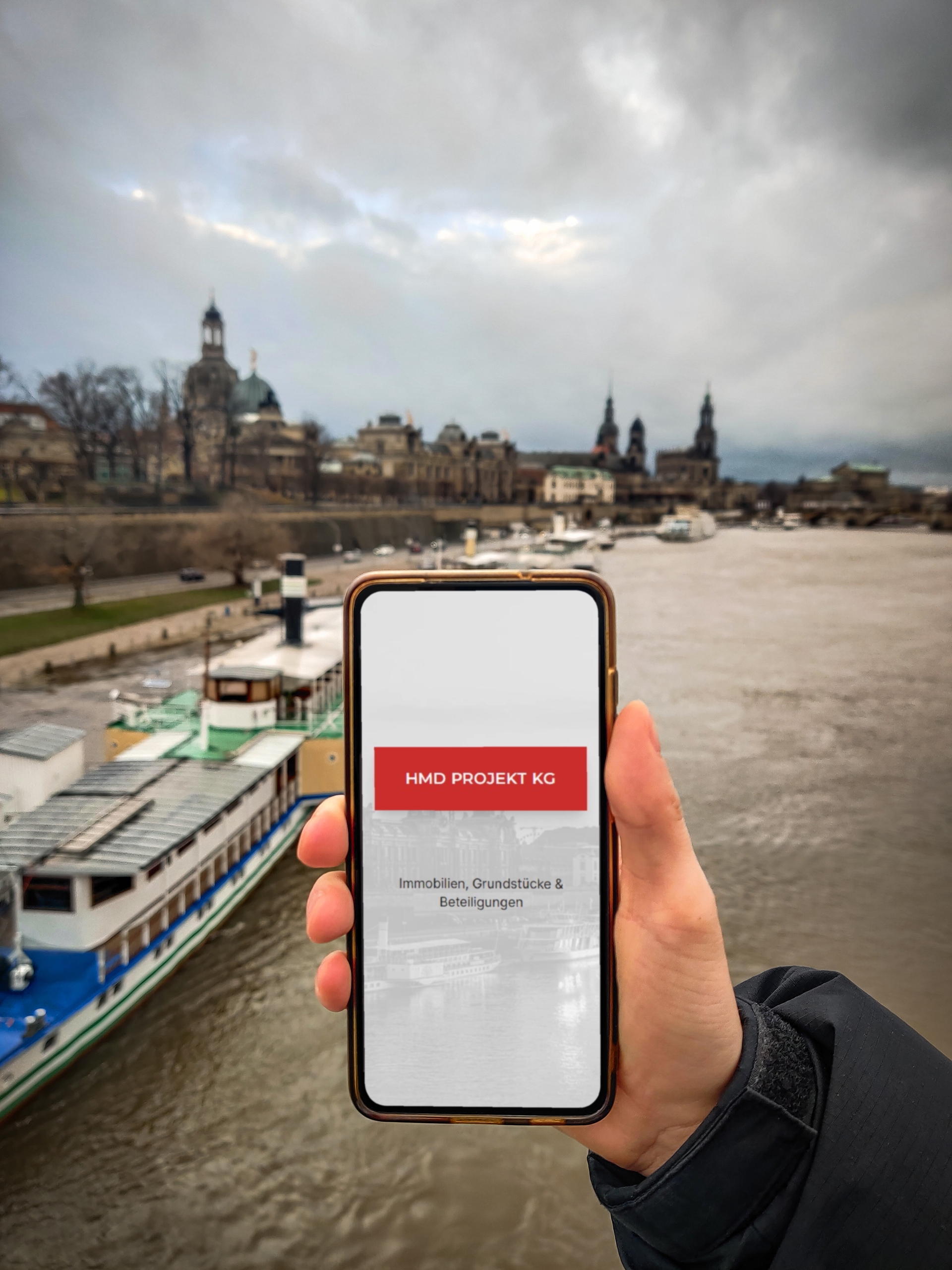 Fotografie eines Smartphones, mit geöffneter hmd.immo Webseite.  Elbe und Stadtsilhouette von Dresden sind im Hintergrund zu sehen.
